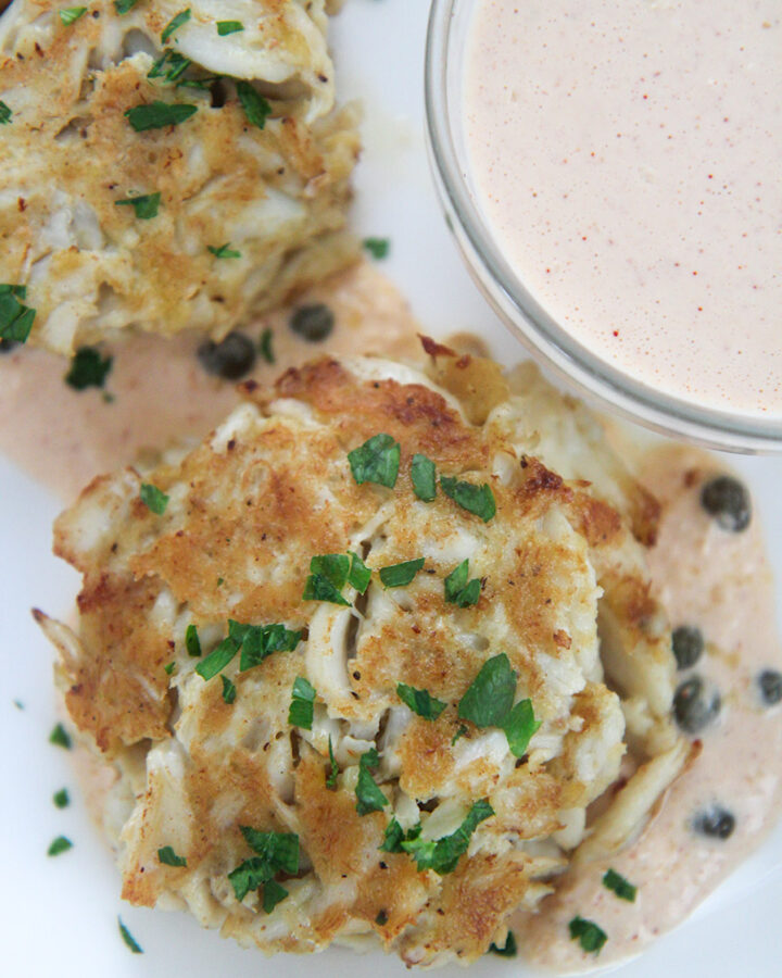two lump crab cakes with parsley on top and a side of remoulade in a small bowl.