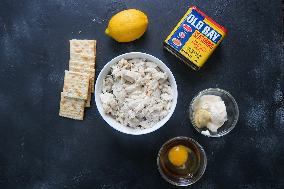 a small bowl of crab meat, a lemon, saltine crackers, old bay seasoning, an egg in a small bowl, and mayo and mustard in a small bowl. 