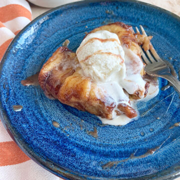 apple dumplings with vanilla ice cream, caramel syrup, and a fork on a blue plate with an orange towel on the side.