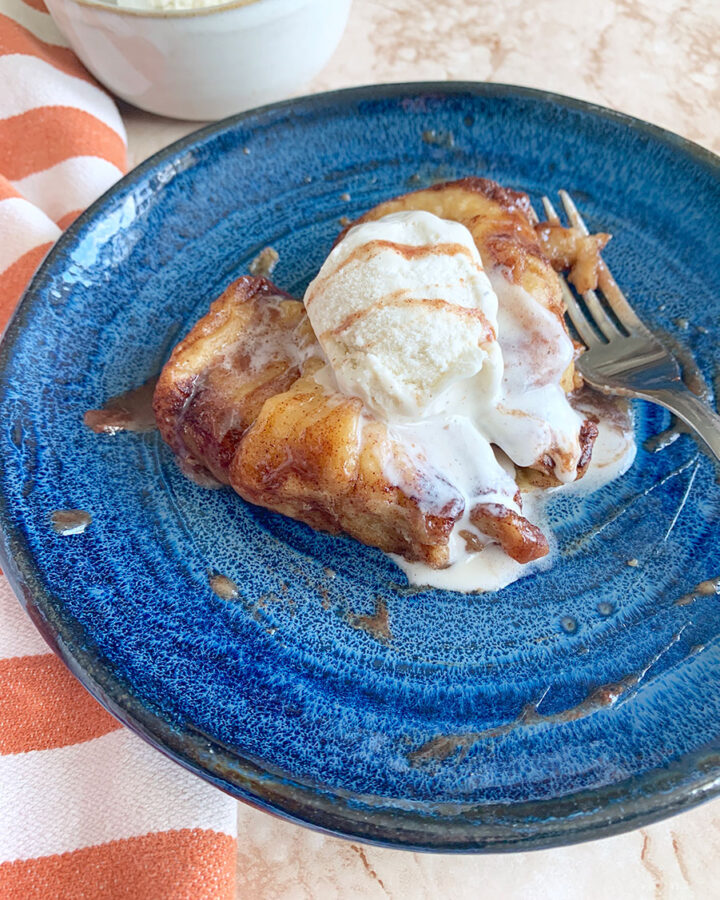 apple dumplings with vanilla ice cream, caramel syrup, and a fork on a blue plate with an orange towel on the side.