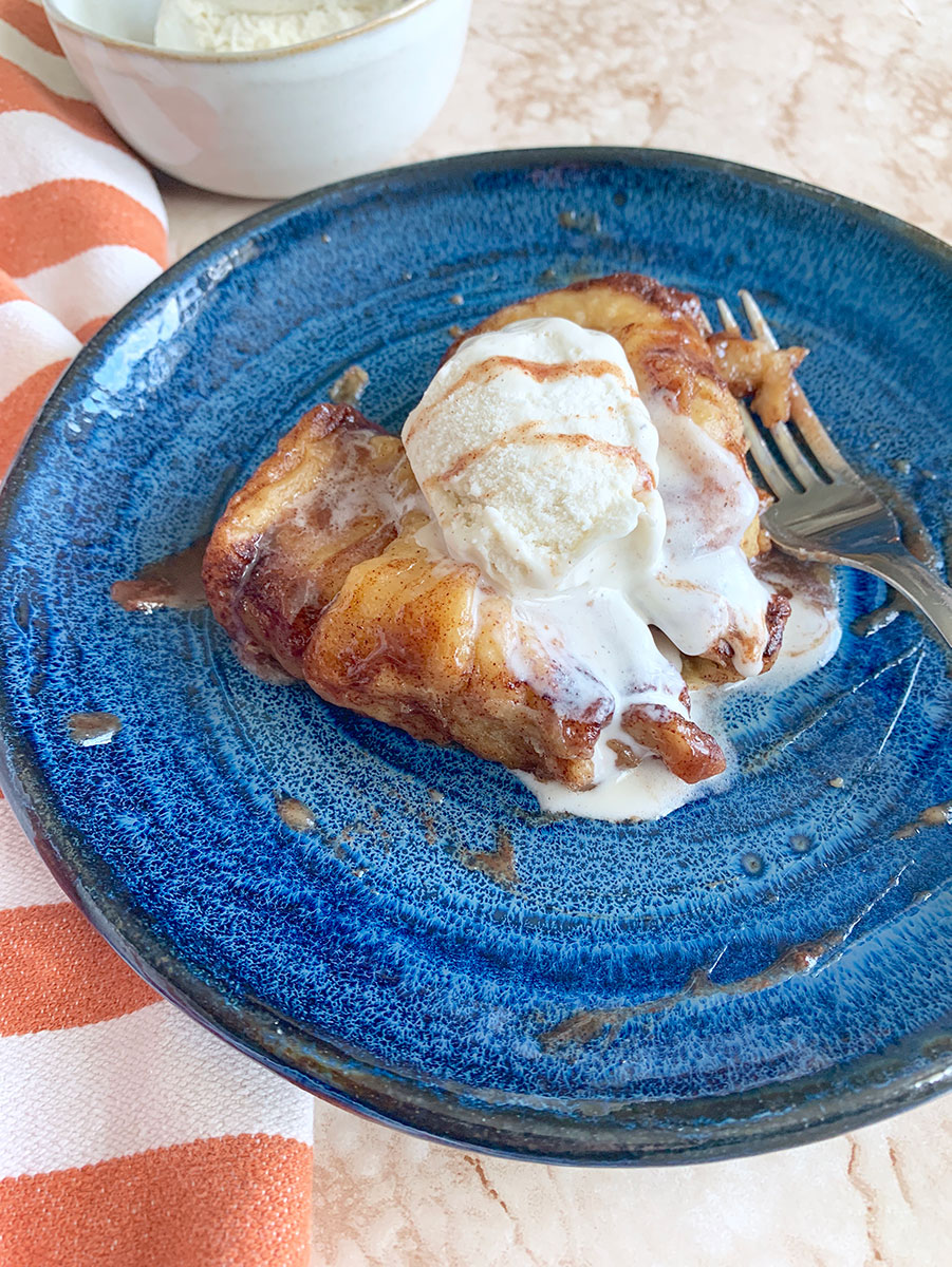 apple dumplings with vanilla ice cream, caramel syrup, and a fork on a blue plate with an orange towel on the side. 