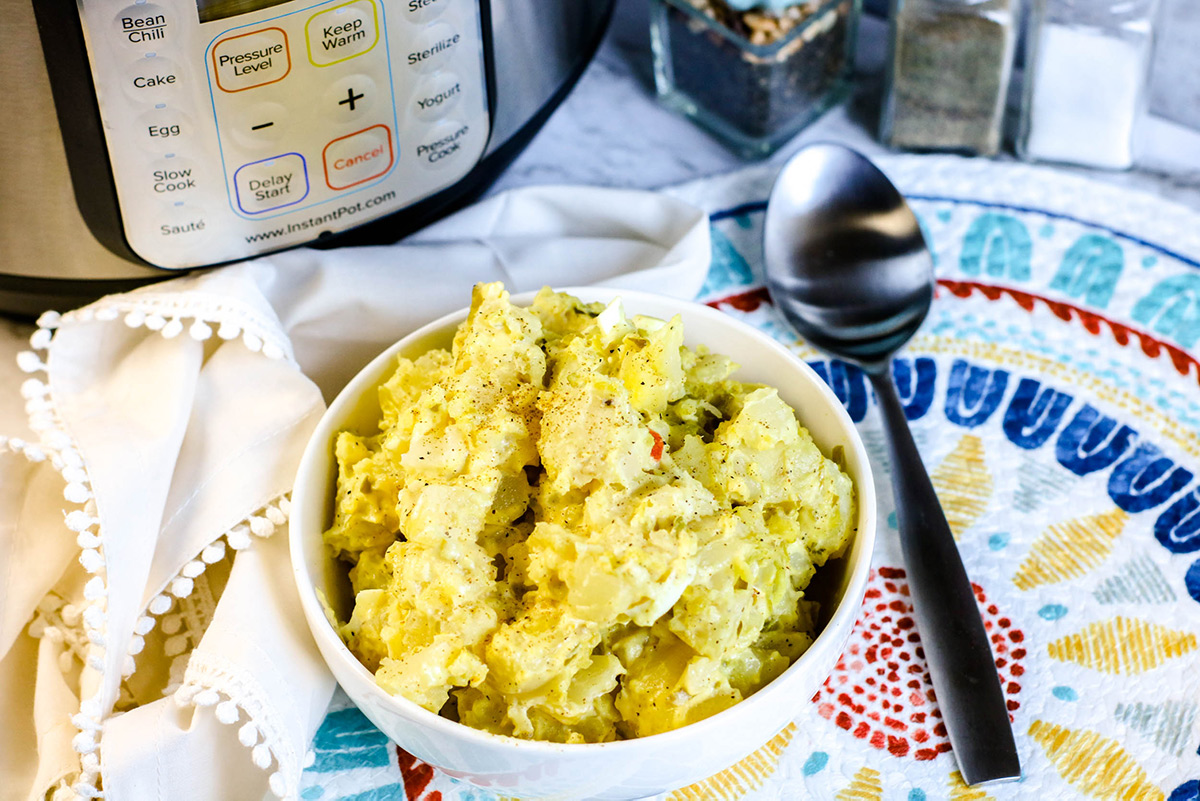 A horizontal photo of a small bowl filled with potato salad, a spoon on the side, an instant pot in the background, and some salt shakers. 