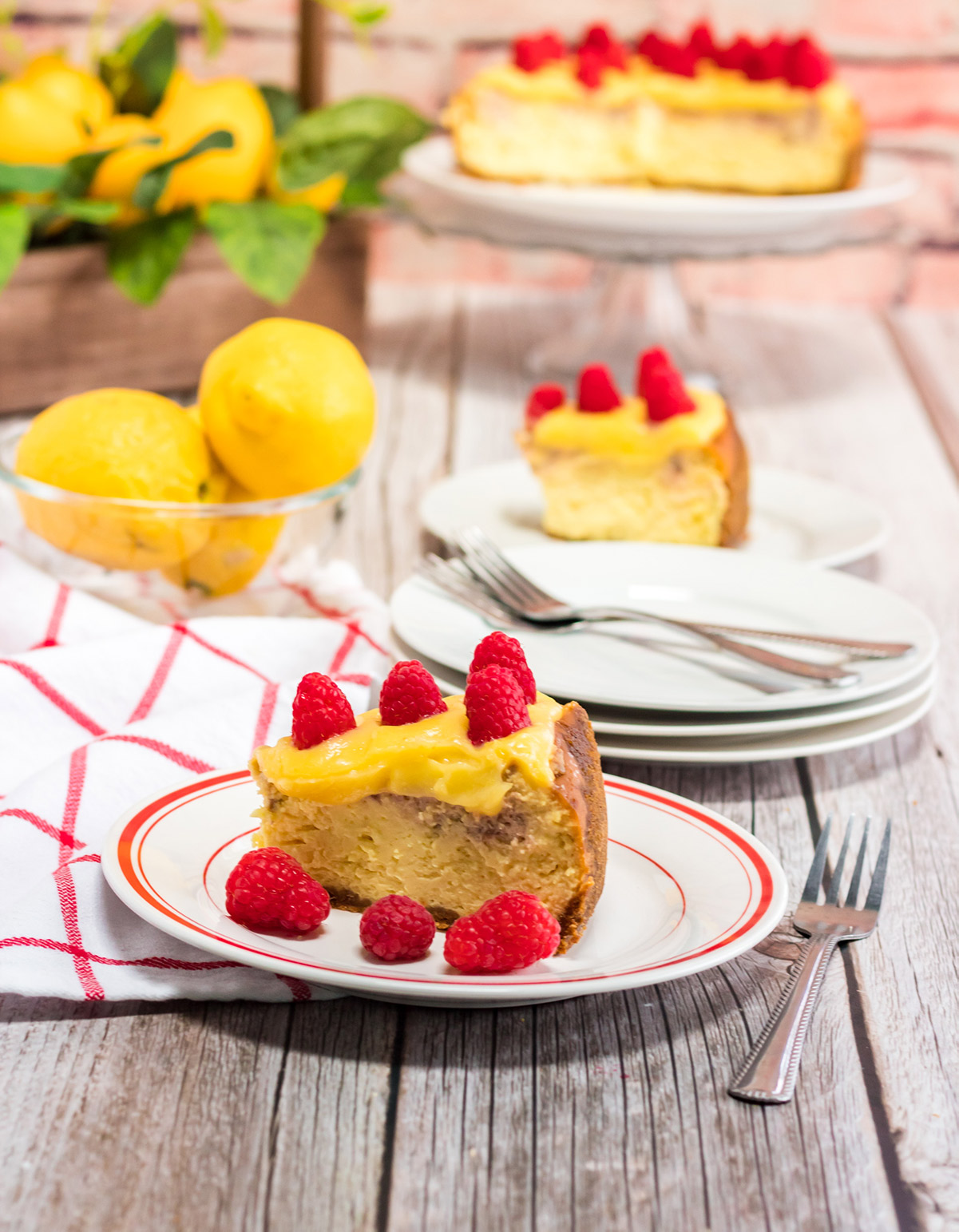 a slice of instant pot raspberry lemonade cheesecake on a white and red plate with a fork, towel, and lemons on the side. 