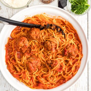 spaghetti and meatballs with a serving spoon in a white bowl with crushed red pepper, parmesan cheese, and fresh basil on the side.