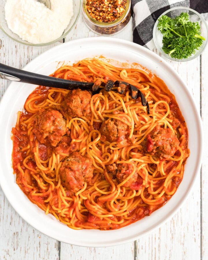 spaghetti and meatballs with a serving spoon in a white bowl with crushed red pepper, parmesan cheese, and fresh basil on the side.