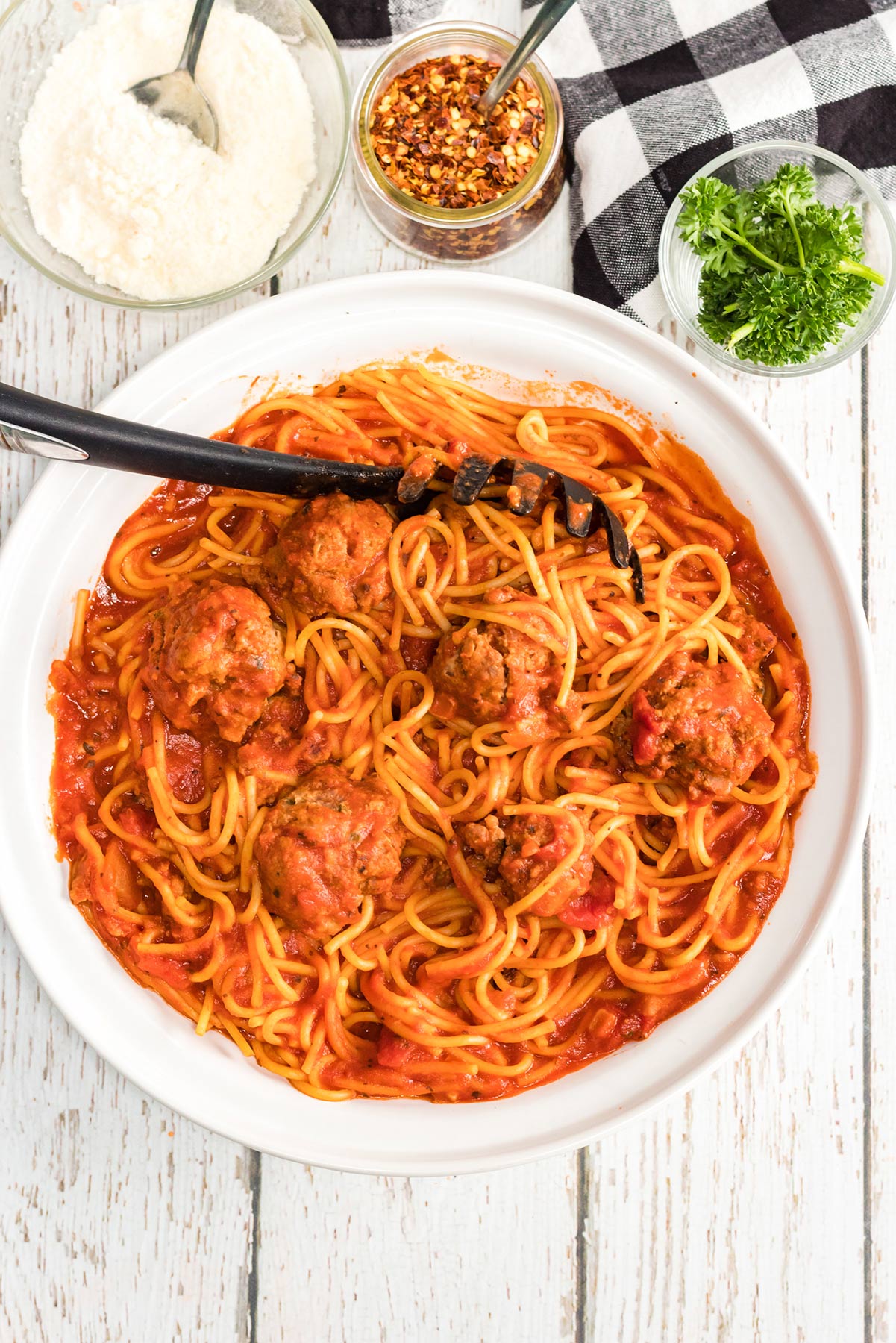 spaghetti and meatballs with a serving spoon in a white bowl with crushed red pepper, parmesan cheese, and fresh basil on the side. 