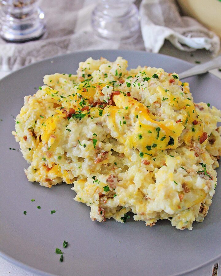 loaded cauliflower bake on a gray plate with salt shakers on the side and a green casserole in the background.