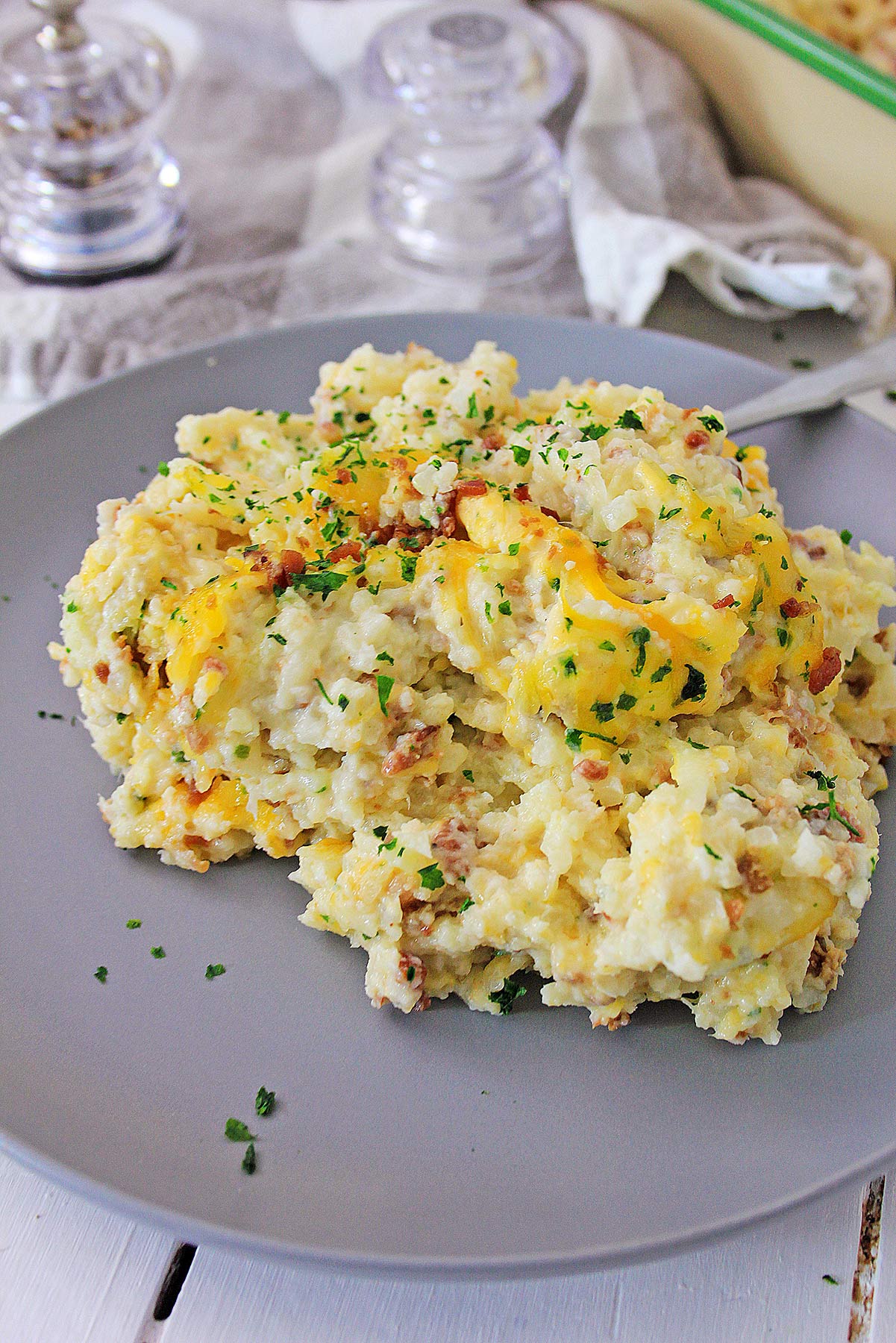 loaded cauliflower bake on a gray plate with salt shakers on the side and a green casserole in the background. 