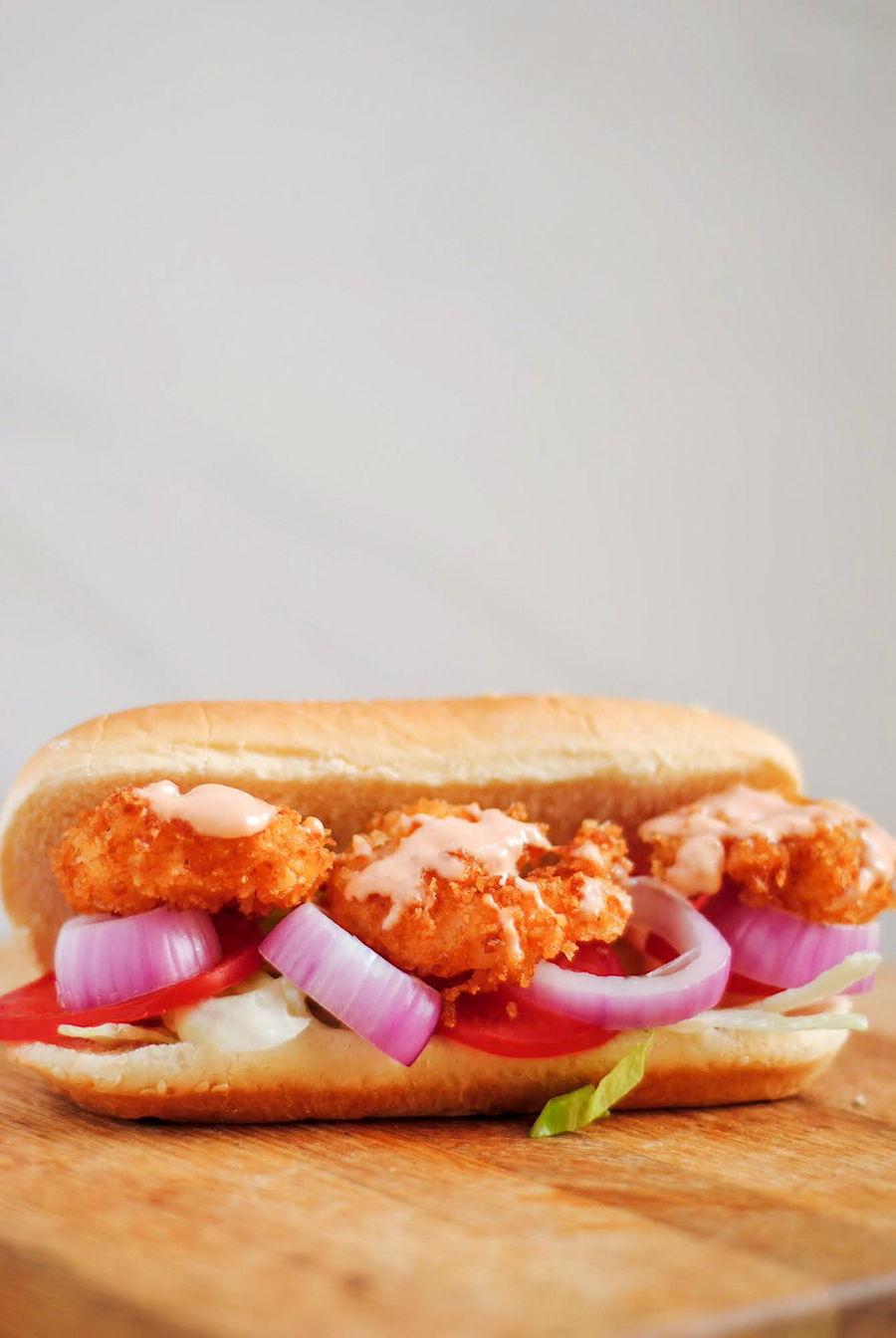 shrimp po'boy sandwich with red onions, lettuce, tomato, and sauce on a wooden board