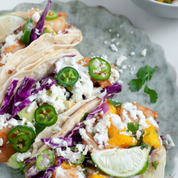 Three shrimp tacos with mango salsa and cheese on top of a light blue plate. A small white bowl of mango salsa on the side.