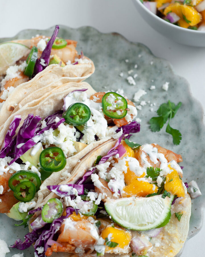 Three shrimp tacos with mango salsa and cheese on top of a light blue plate. A small white bowl of mango salsa on the side.