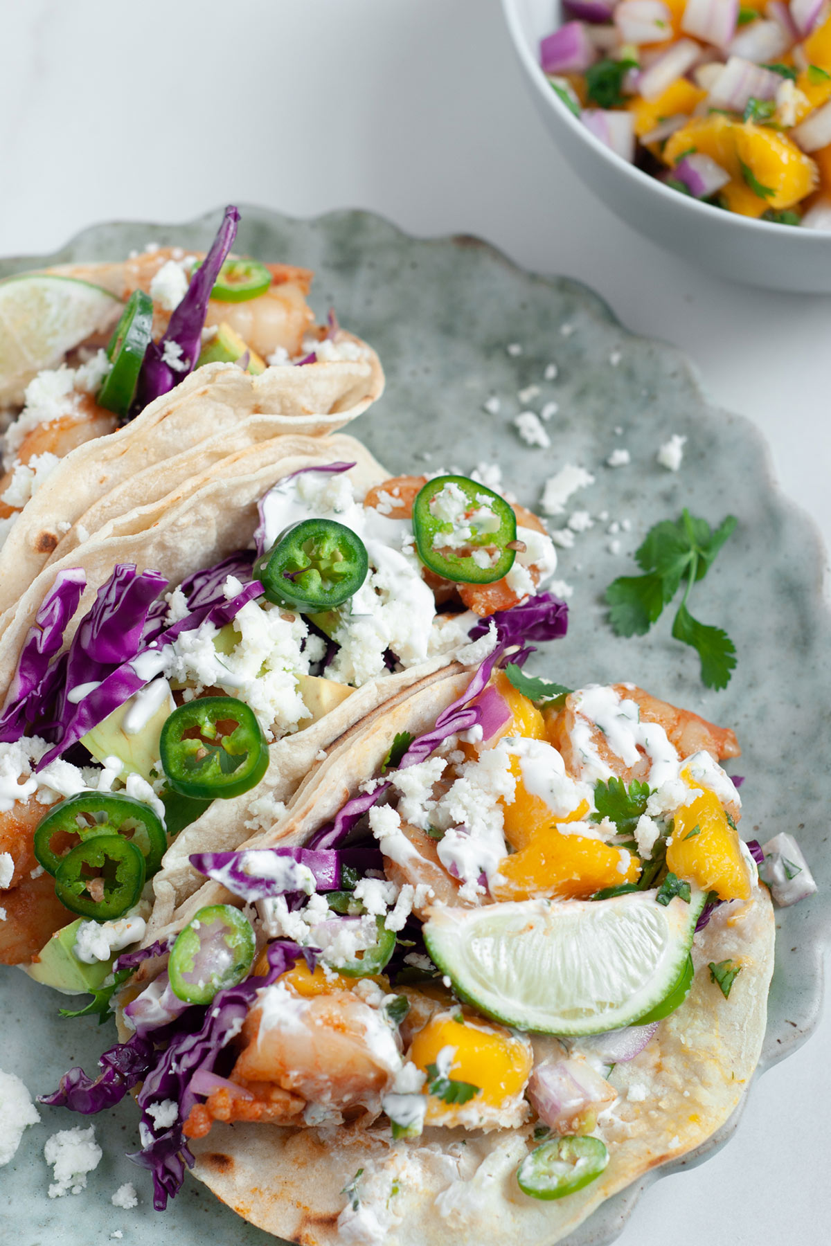 Three shrimp tacos with mango salsa and cheese on top of a light blue plate. A small white bowl of mango salsa on the side. 