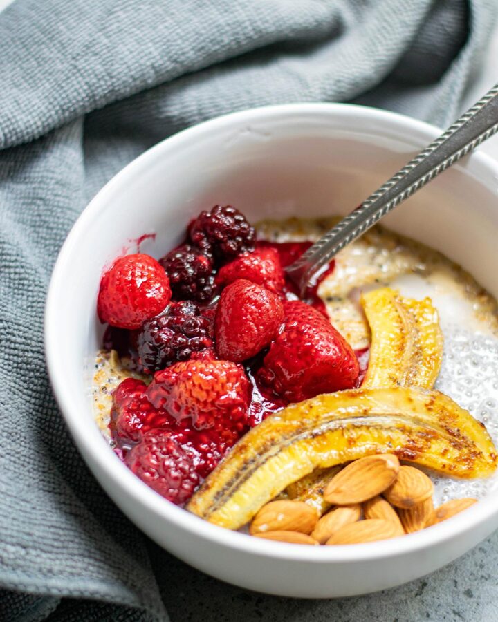 a white bowl with oats, berries, bananas, nuts, and a spoon. A blue towel on the side.