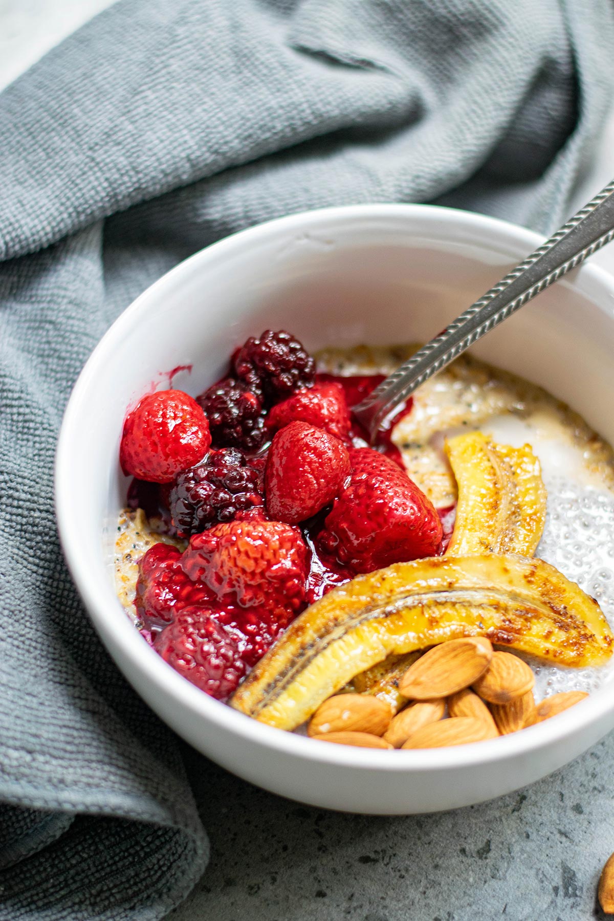 a white bowl with oats, berries, bananas, nuts, and a spoon. A blue towel on the side. 