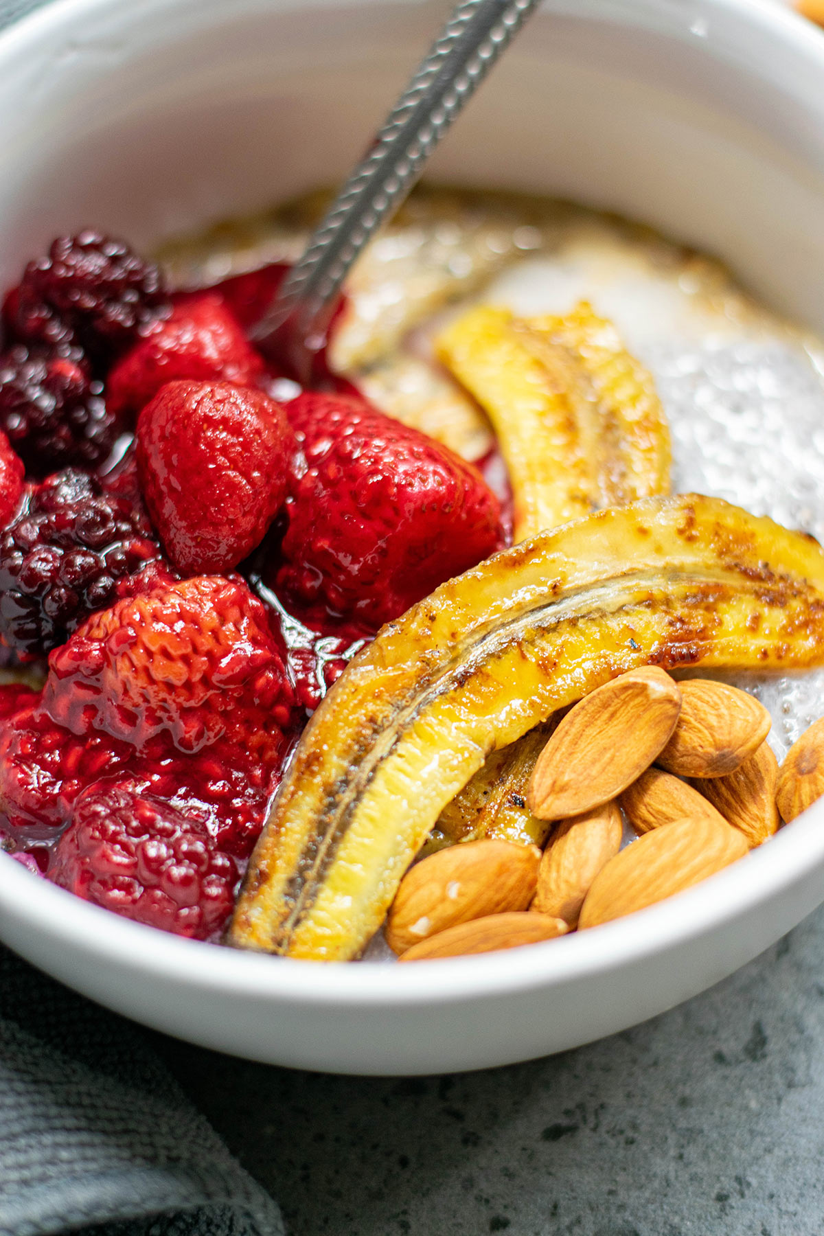 a close up shot of mixed berry and banana oats with nuts and a spoon in a white bowl. 