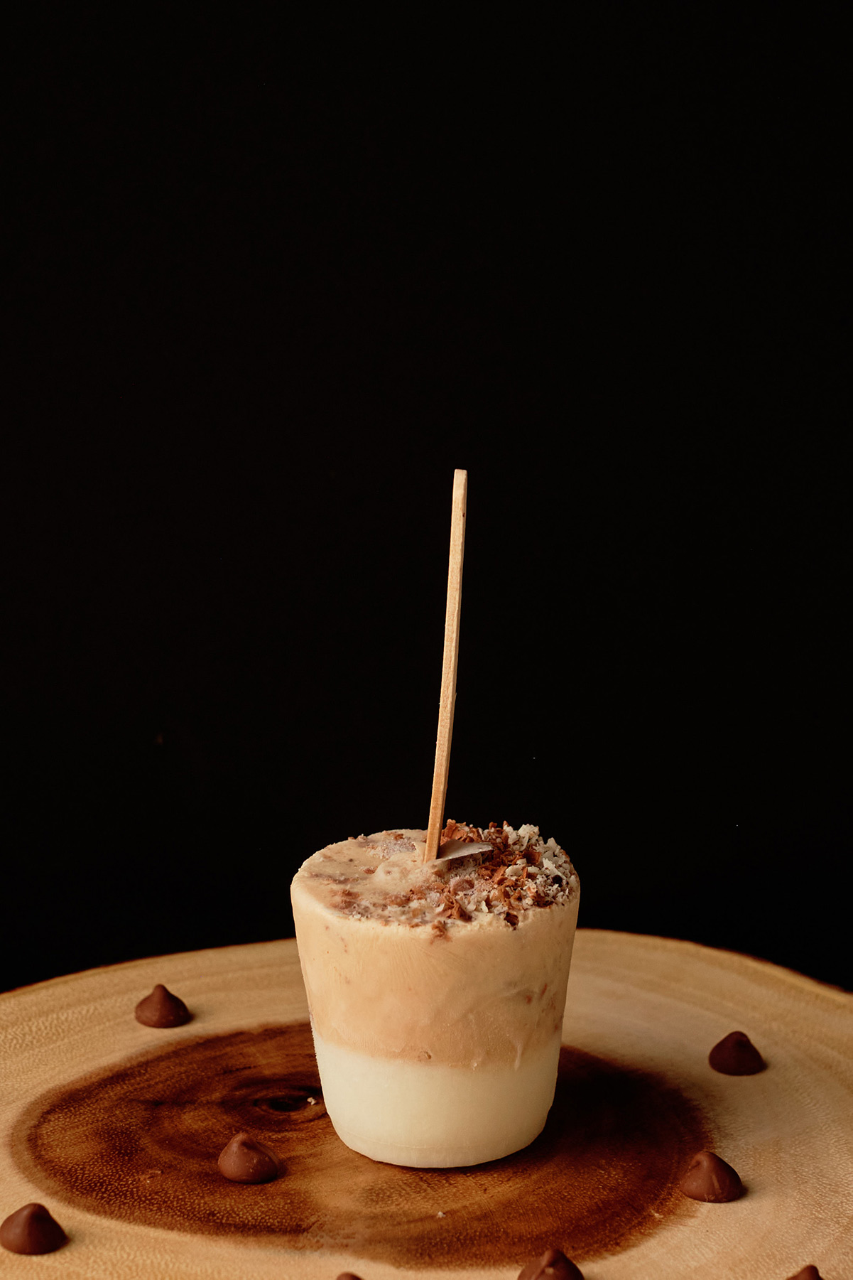 white chocolate peanut butter popsicle on a wooden circular board with chocolate chips on the side and a black backdrop. 