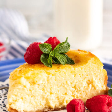 a slice of creme brulee cheesecake up close with raspberries, mint, and a glass of milk in the background.