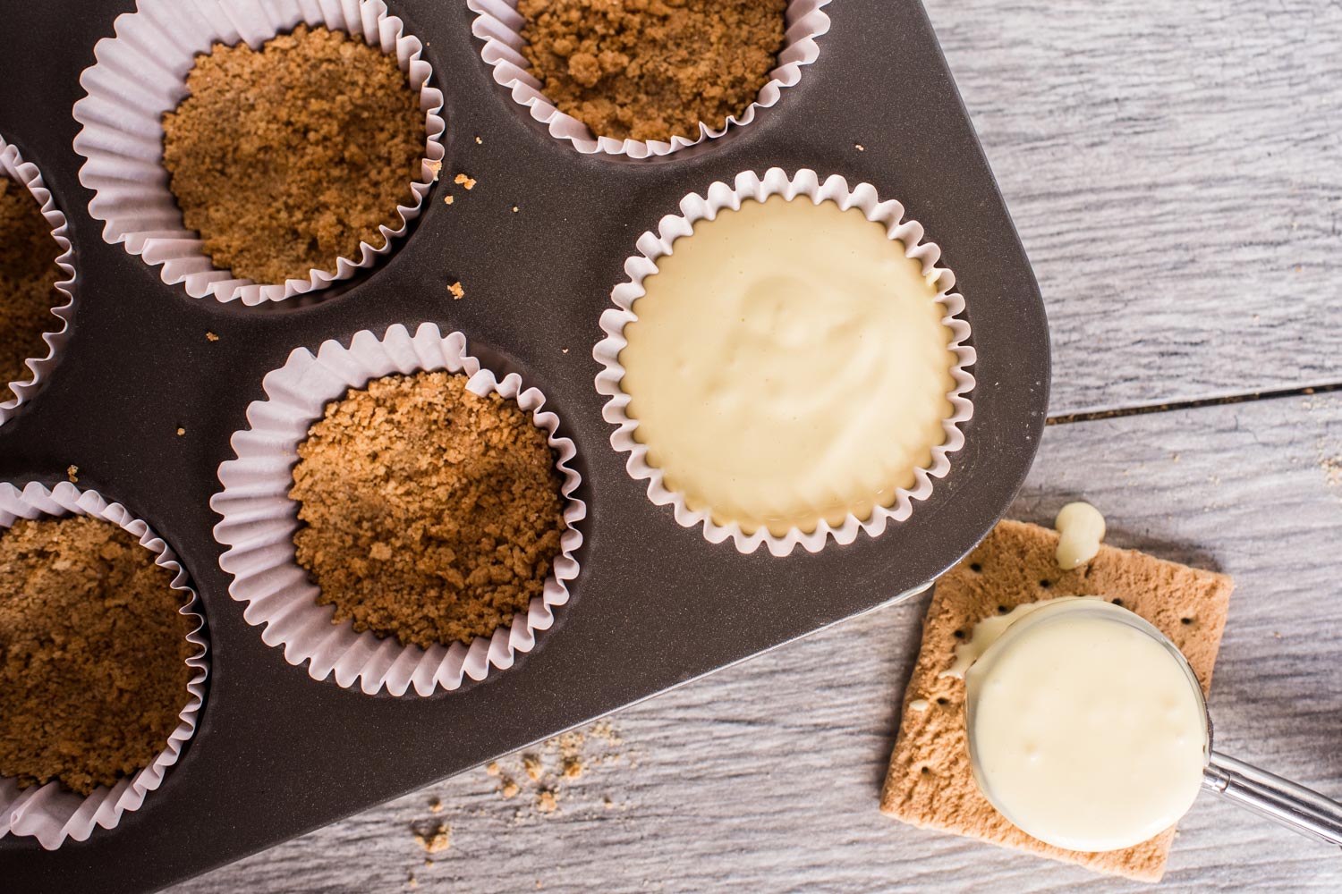 cupcake tin with cupcake liners filled with graham cracker crust and cheesecake filling.