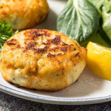 Two crab cakes on a plate with a lemon wedge and salad.