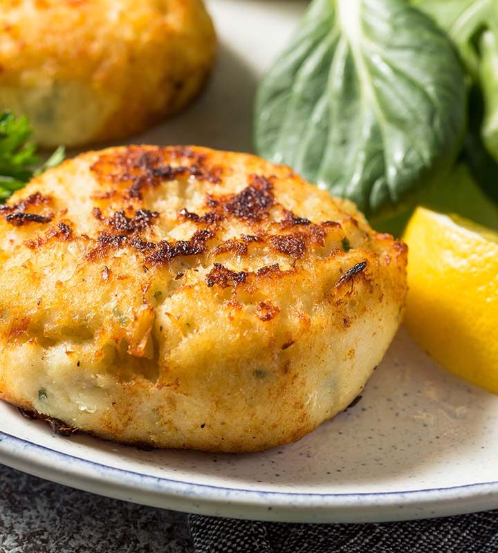 Two crab cakes on a plate with a lemon wedge and salad.