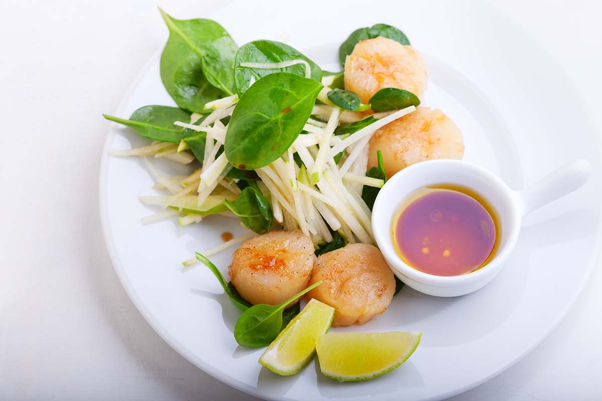 cooked bay scallops on a white plate with a lime wedge and sauce on the side.