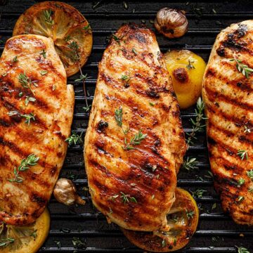 Three grilled chicken breasts on a grill with lemons, herbs, and garlic.