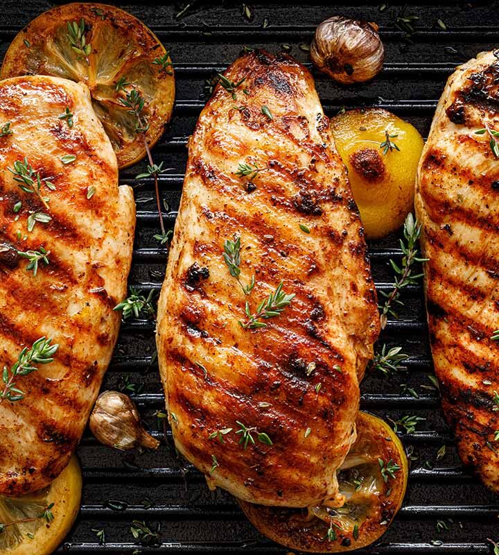 Three grilled chicken breasts on a grill with lemons, herbs, and garlic.