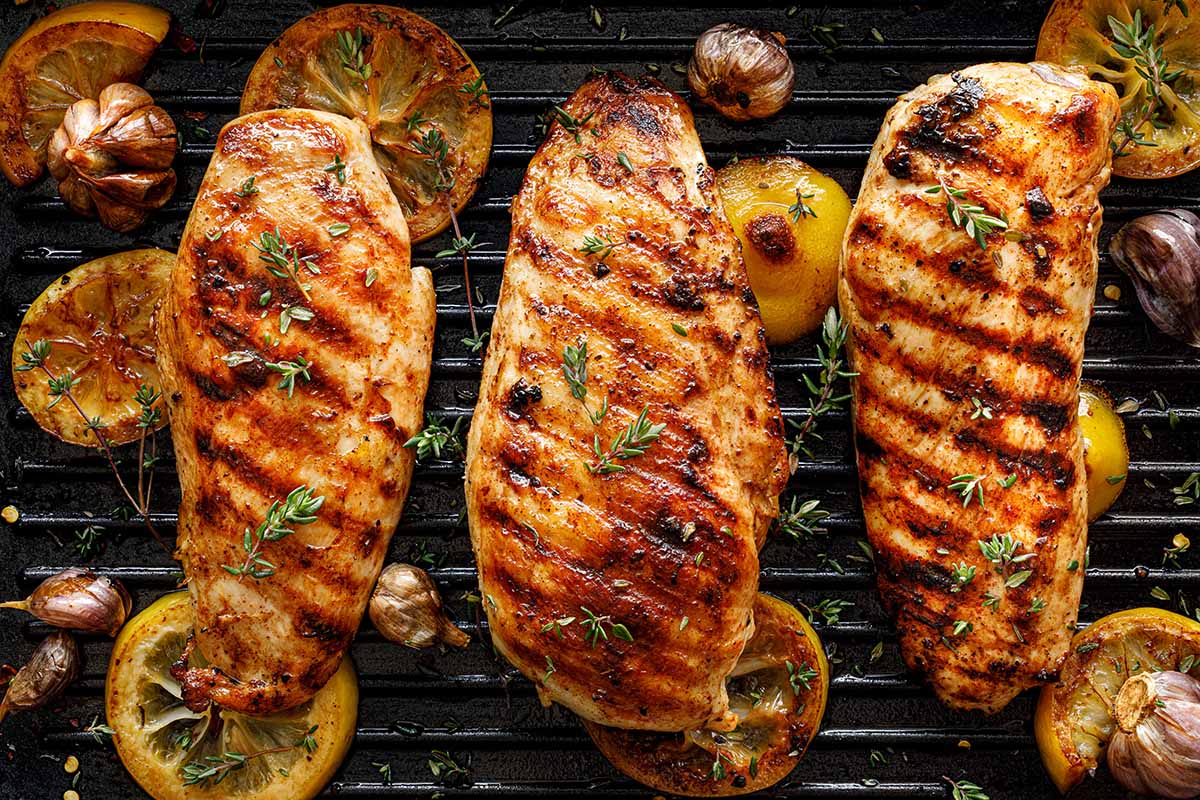 Three grilled chicken breasts on a grill with lemons, herbs, and garlic. 