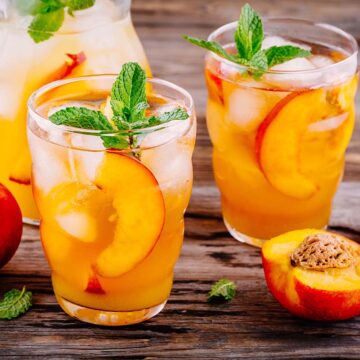 two glasses of peach crown royal lemonade and a jug and peaches in the background.