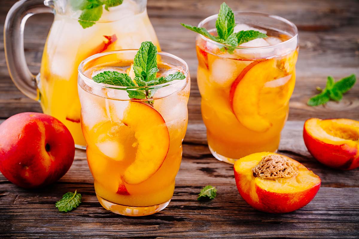 two glasses of peach crown royal lemonade and a jug and peaches in the background.